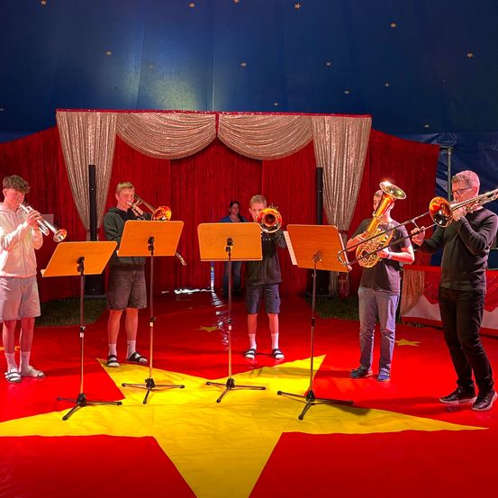 Eine Gruppe von Musikern steht auf einem roten Boden mit einem gelben Stern, trägt Freizeitkleidung und hält Posaunen und Trompeten. Ein roter Vorhang steht hinter ihnen, und eine Person steht hinten.