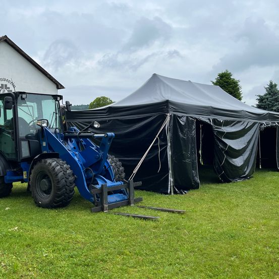 Ein blauer Traktor steht neben einem großen schwarzen Zelt auf einem Rasenfeld, mit einem Haus und einem bewölkten Himmel im Hintergrund.