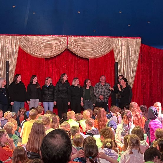 Eine Gruppe von Erwachsenen und Kindern versammelt sich unter einem Zelt mit roten und goldenen Vorhängen. Einige Erwachsene stehen auf der Bühne, während Kinder im Publikum sitzen und verschiedene Jacken und Rucksäcke tragen.