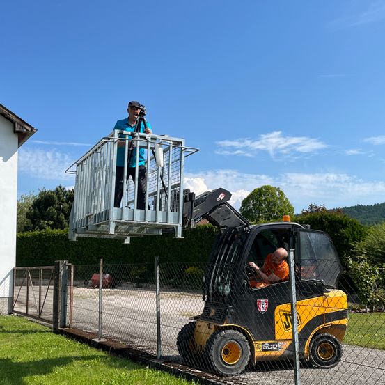 Ein Mann mit einer Kamera steht auf einer Hebebühne auf einem Gabelstapler, vor einem Haus und einem Zaun positioniert, unter einem klaren Himmel.