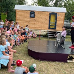 Eine Gruppe von Kindern und Erwachsenen beobachtet einen Hund auf der Bühne vor einem kleinen Haus. Der Hund steht hinter einem Geländer.