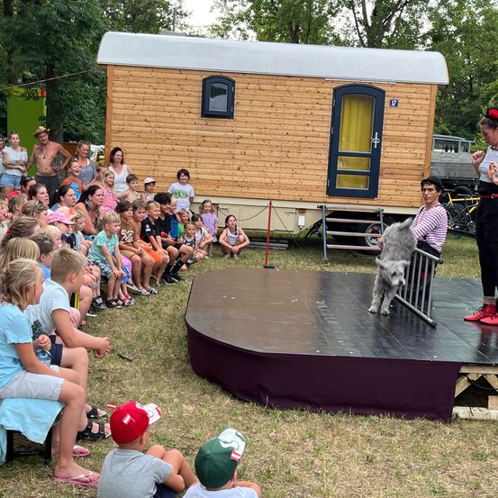 Eine Gruppe von Kindern und Erwachsenen beobachtet einen Hund auf der Bühne vor einem kleinen Haus. Der Hund steht hinter einem Geländer.