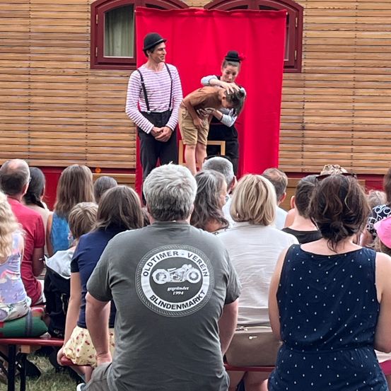 Eine Straßenaufführung mit einem Mann und einer Frau, eine rückwärts gebeugt, vor einem Publikum. Ein Mann im Publikum trägt ein graues Shirt mit Logo.