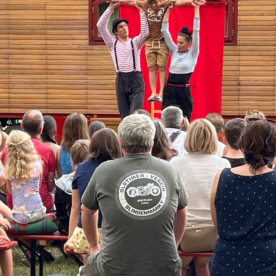 Eine Menschenmenge beobachtet eine Vorstellung von einem Mann, einer Frau und einem Kind auf einer hölzernen Bühne. Der Mann und die Frau balancieren das Kind auf ihren Händen.