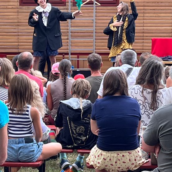 Zwei Darsteller unterhalten ein Publikum auf der Bühne. Einer hält ein grünes Objekt und der andere hebt die Arme. Das Publikum sitzt auf Bänken und schaut zu.