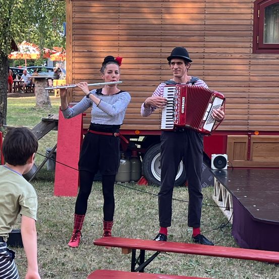 Zwei Künstler stehen auf einer Bühne mit einem hölzernen Hintergrund. Eine Frau spielt Flöte, während ein Mann Akkordeon spielt. Ein Kind beobachtet sie vom Gras.