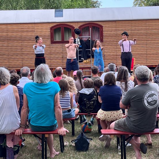 Eine Gruppe von Darstellern auf einer Bühne vor einem hölzernen Wohnwagen, mit einem Publikum, das zuschaut. Die Darsteller tragen Freizeitkleidung und scheinen mitten in einer Aufführung zu sein.