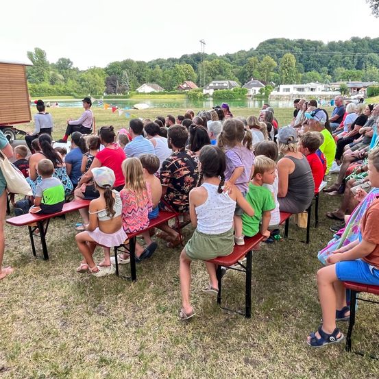 Eine große Gruppe von Menschen, einschließlich Kindern und Erwachsenen, sitzen auf Bänken rund um einen See und hören einem Sprecher zu. Eine Person steht auf einer Bühne mit einer Kulisse aus Bäumen.