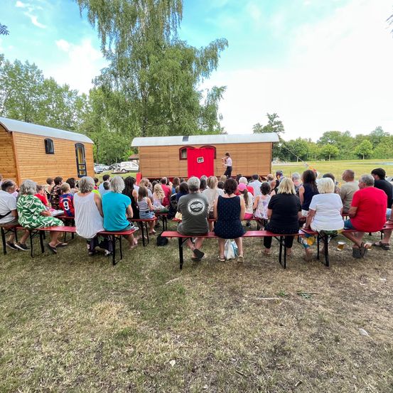 Eine Gruppe von Menschen sitzt auf Bänken um ein kleines Holzhaus mit einer roten Tür. Eine Person steht vor dem Haus, vielleicht auf der Bühne. Bäume und ein grasiger Feld im Hintergrund.