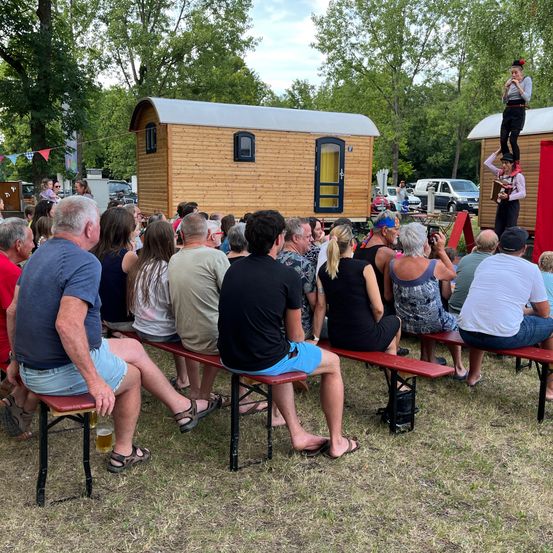 Eine Menschenmenge sitzt auf Bänken und beobachtet einen Künstler in einem kleinen Holzhaus auf Stelzen. Bäume und geparkte Autos sind im Hintergrund.