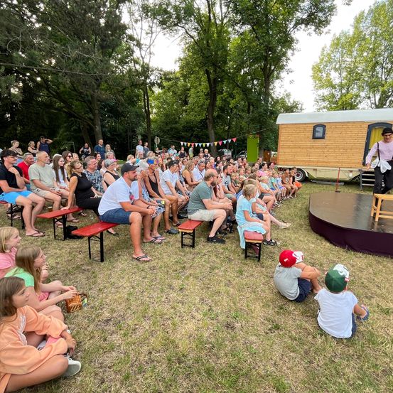 Eine Gruppe von Menschen unterschiedlichen Alters ist in einem Außenbereich, vielleicht einem Park, versammelt, um eine Aufführung zu sehen. Ein Künstler steht mit einem kleinen Wohnwagen hinter ihm auf der Bühne. Das Publikum sitzt auf Bänken und auf der Wiese.