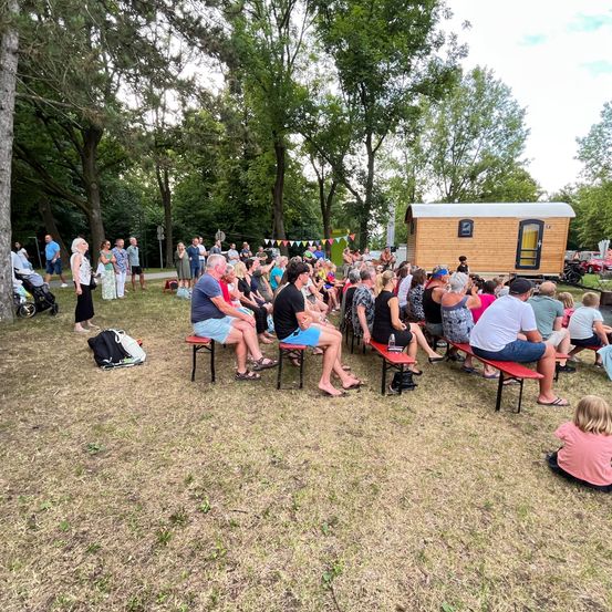 In einem Park versammelt sich eine Menschenmenge um ein Tiny House und lauscht aufmerksam. Einige sitzen auf Bänken, andere stehen. Bunte Fahnen hängen im Hintergrund, und ein Kinderwagen ist in der Nähe. Bäume spenden Schatten.