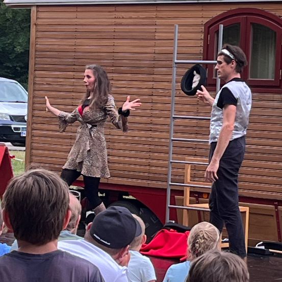 Eine Frau und ein Mann führen ein Straßenkunststück vor einem Holzhaus auf. Die Frau hat ihre Arme ausgestreckt, während der Mann neben einer Leiter steht. Ein Publikum beobachtet von unten.
