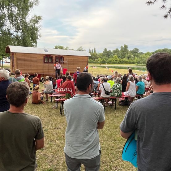 Eine Gruppe von Menschen versammelt sich auf einem Feld und sieht sich eine Aufführung auf einer Bühne an. Die Bühne ist ein kleines Holzhaus mit einer Leiter. Einige Leute sitzen auf Bänken, während andere stehen. Bäume und ein Gewässer sind im Hintergrund.