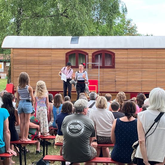 Zwei Darsteller stehen vor einem hölzernen Wohnwagen, während sich ein Publikum unterschiedlichen Alters versammelt, um zuzuschauen. Der Wohnwagen hat zwei Fenster, und die Darsteller halten Papiere. Bäume und ein Gebäude sind im Hintergrund.