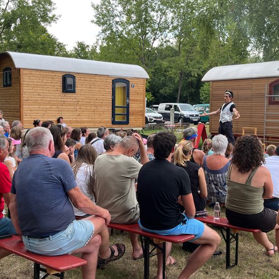 Eine Menschenmenge sitzt auf Bänken vor zwei Holzhütten und beobachtet einen Künstler auf der Bühne. Die Hütten haben geneigte Dächer und sind von Bäumen umgeben.
