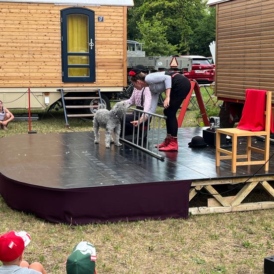 Eine Frau in roten Stiefeln steht mit einem Hund auf einer Bühne. Ein Holzstuhl und ein roter Stoff sind auf der Bühne. Eine Frau und ein Kind sitzen auf dem Boden, und ein Anhänger steht dahinter.