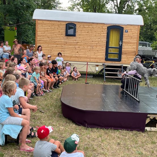 Eine Menschenmenge hat sich versammelt, um eine Vorstellung auf einer Bühne mit einem Hund zu sehen. Die Bühne ist von einem kleinen Holzhaus mit einem kleinen Fenster und einer Tür umgeben.