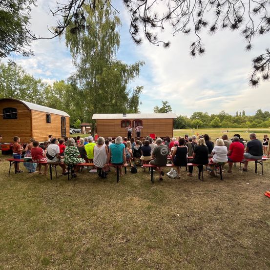 Eine Menschenmenge versammelt sich im Freien für eine Aufführung vor zwei kleinen Holzhütten unter einem teilweise bewölkten Himmel.