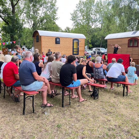 Eine Gruppe von Menschen sitzt auf Bänken vor einer hölzernen Bühne und schaut sich eine Aufführung an. Hinter ihnen stehen zwei kleine Hütten.