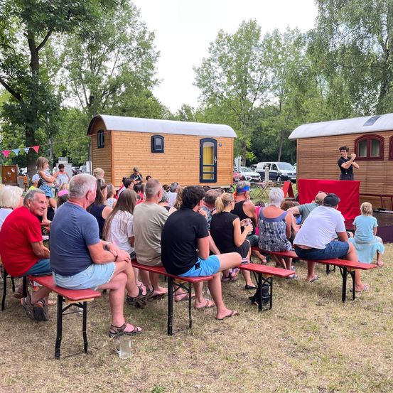 Eine Gruppe von Menschen sitzt auf Bänken in einem Außenbereich, vielleicht für eine Veranstaltung. Vor ihnen steht eine Person und spricht auf einer Bühne. Dahinter befinden sich zwei winzige Häuser und ein roter Stoff, der an der Bühne hängt.