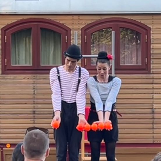 Zwei Künstler in traditioneller Clown-Kleidung jonglieren mit orangefarbenen Bällen vor einem Holzgebäude. Ein Mensch beobachtet von hinten.