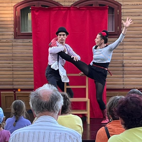 Ein Mann und eine Frau führen akrobatische Darbietungen auf einer Bühne vor einem Publikum auf, mit einem roten Vorhang als Hintergrund.