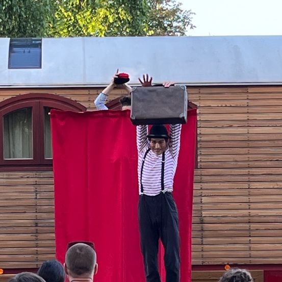 Ein Mann führt einen Balanceakt mit einer Tasche auf dem Kopf vor einem Holzgebäude durch. Das Publikum beobachtet ihn von unten.