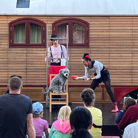 Zwei Künstler stehen auf der Bühne mit einem Hund vor einer Holzwand. Einer steht auf einer Leiter mit einem Hund, während der andere einen Ball hält. Ein Publikum beobachtet von vorne.