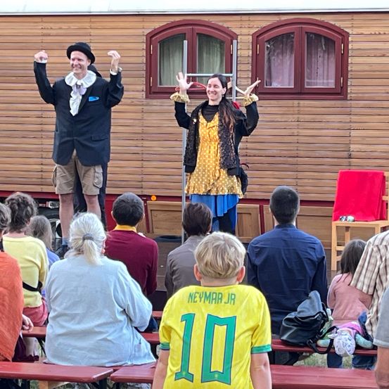Zwei Personen treten vor einem Holzhaus auf der Bühne auf. Das Publikum sitzt auf Bänken vor der Bühne. Ein Kind in einem gelben Trikot mit der Nummer 10 schaut zu.