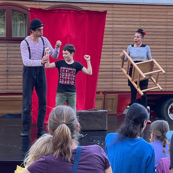 Eine Gruppe von Darstellern auf der Bühne, einer hält einen Stuhl, ein anderer interagiert mit einem Jungen, während ein Publikum zuschaut.