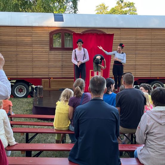 Eine Zirkusvorstellung findet vor einem hölzernen Wagen statt, während eine Menge zuschaut. Performer in Kostümen sind auf der Bühne, einer macht einen Handstand und eine andere reicht aus. Das Publikum sitzt auf roten Bänken.