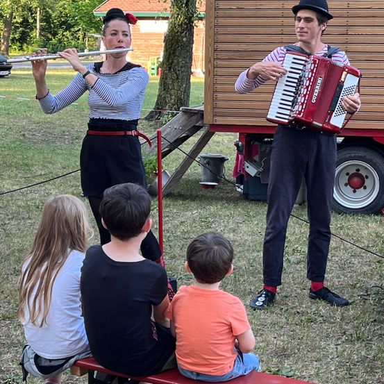 Eine Frau spielt Flöte, während ein Mann Akkordeon spielt. Mehrere Kinder sitzen auf einer Bank und beobachten sie. Bäume, ein Haus und ein Fahrzeug sind im Hintergrund.