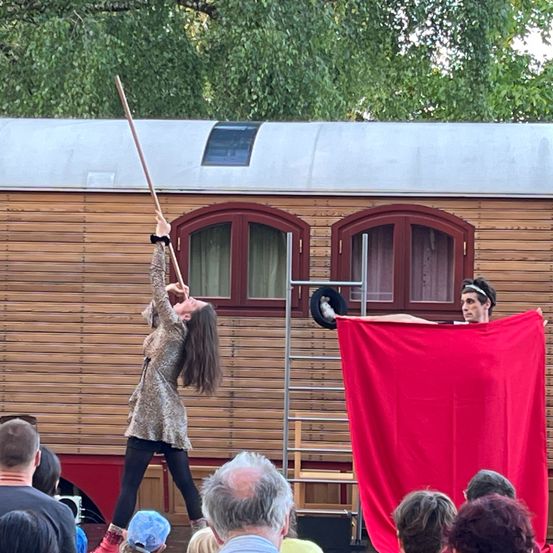 Ein Darsteller hält einen langen Stock, während ein anderer auf einem roten Tuch liegt, beobachtet von einem Publikum vor einem Holzgebäude.