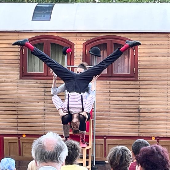 Ein Mann führt einen akrobatischen Handstand auf einem Stuhl aus, während ein anderer ihn aufrecht hält. Sie stehen vor einem Gebäude mit Holzwänden, das von Zuschauern beobachtet wird.