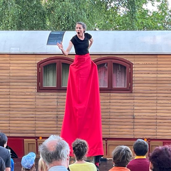 Eine Frau in einem roten Rock balanciert auf Stelzen vor einem hölzernen Wagen, während eine Menge zuschaut.