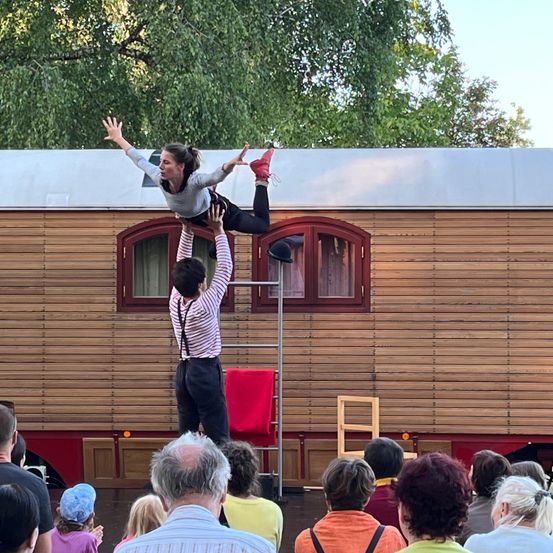 Ein Zirkusauftritt im Freien, bei dem eine Frau auf den Händen eines Mannes balanciert, während eine Menge zuschaut. Die Umgebung besteht aus einem hausartigen, hölzernen Gebäude mit Fenstern und einem roten Tuch.