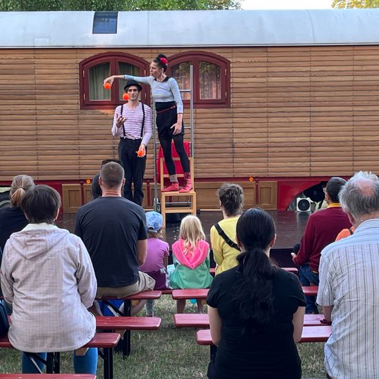 Zwei Künstler unterhalten ein Publikum vor einem hölzernen Wohnwagen. Eine Frau balanciert auf einer Leiter und jongliert, während ein Mann darunter steht. Das Publikum beobachtet das Treiben von Bänken aus.