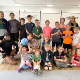 Eine Gruppe von Kindern und Erwachsenen posiert für ein Foto in einem Fitnessstudio. Die Kinder tragen Medaillen und halten einen blauen Ball.