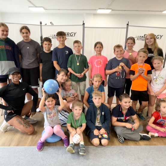 Eine Gruppe von Kindern und Erwachsenen posiert für ein Foto in einem Fitnessstudio. Die Kinder tragen Medaillen und halten einen blauen Ball.