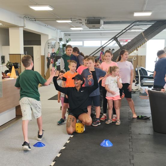 Eine Gruppe von Kindern und einer erwachsenen Frau in einem Fitnessstudio. Die Frau kniet mit erhobenen Händen, während die Kinder zusehen. Einige stehen hinter ihr, während andere davor sind.