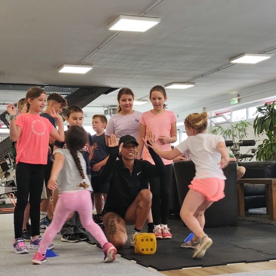 Eine Frau hockt mit einem Würfel in einem Fitnessstudio, während Kinder um sie herum spielen. Sie tragen alle Sportkleidung.