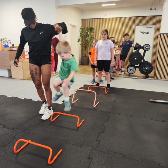 Eine Frau und ein Kind steigen über orange Hürden auf einem Fitnessstudioboden. Dahinter stehen mehrere Kinder und eine Frau. Fitnessgeräte stehen an der Seite.