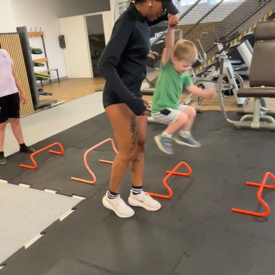 Eine Frau und ein kleiner Junge sind in einem Fitnessstudio. Die Frau hält die Hände des Jungen, während er mit Hürden auf dem Boden springt.