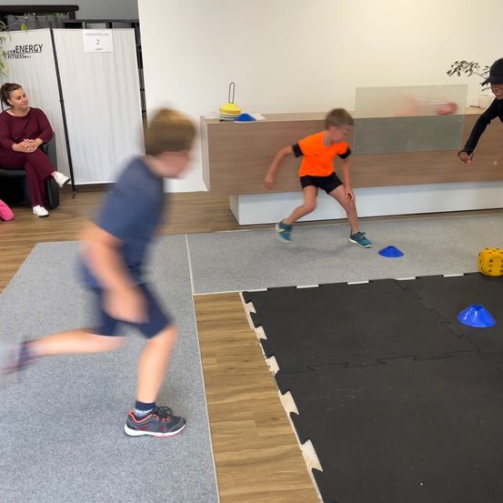 Zwei Jungen spielen in einem Fitnessstudio. Ein Junge läuft, während der andere Junge über Hindernisse springt. Es gibt einen Schreibtisch mit Glasabdeckung. Eine Frau sitzt auf einem Stuhl.