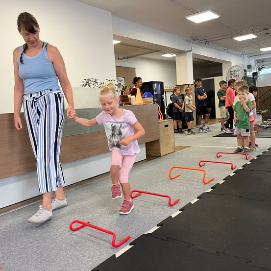 Eine Frau führt ein kleines Mädchen durch eine Hindernisparcours mit roten und orangefarbenen Hürden, während mehrere Kinder zuschauen.