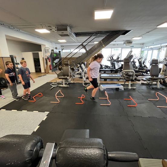 Eine Frau läuft auf einer Matte mit orangefarbenen Hindernissen in einem Fitnessstudio. Zwei Jungen stehen in der Nähe, und verschiedene Fitnessgeräte sind im Hintergrund zu sehen.