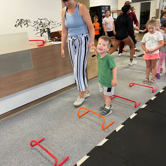 Eine Frau und ein kleiner Junge üben das Springen über Hindernisse in einer Turnhalle. Die Frau hält die Hand des Jungen, während er springt. In der Nähe befinden sich mehrere Kinder und Erwachsene, die verschiedene Aktivitäten ausführen.