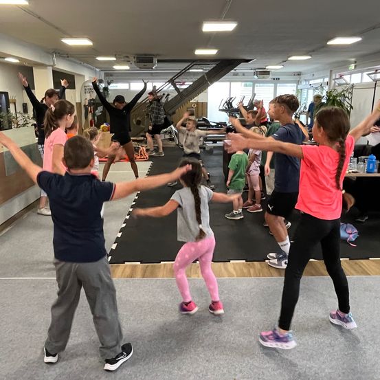 Eine Gruppe von Kindern und Erwachsenen macht Ausfallschritte in einem Fitnessstudio. Einige sind barfuß, andere sind in Turnschuhen. Eine Frau in Schwarz führt sie an.
