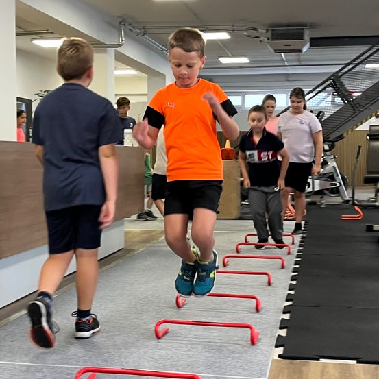 Mehrere Kinder üben Sprungübungen in einem Fitnessstudio. Ein Kind springt über eine Reihe roter Hürden. Dahinter springen auch andere Kinder.